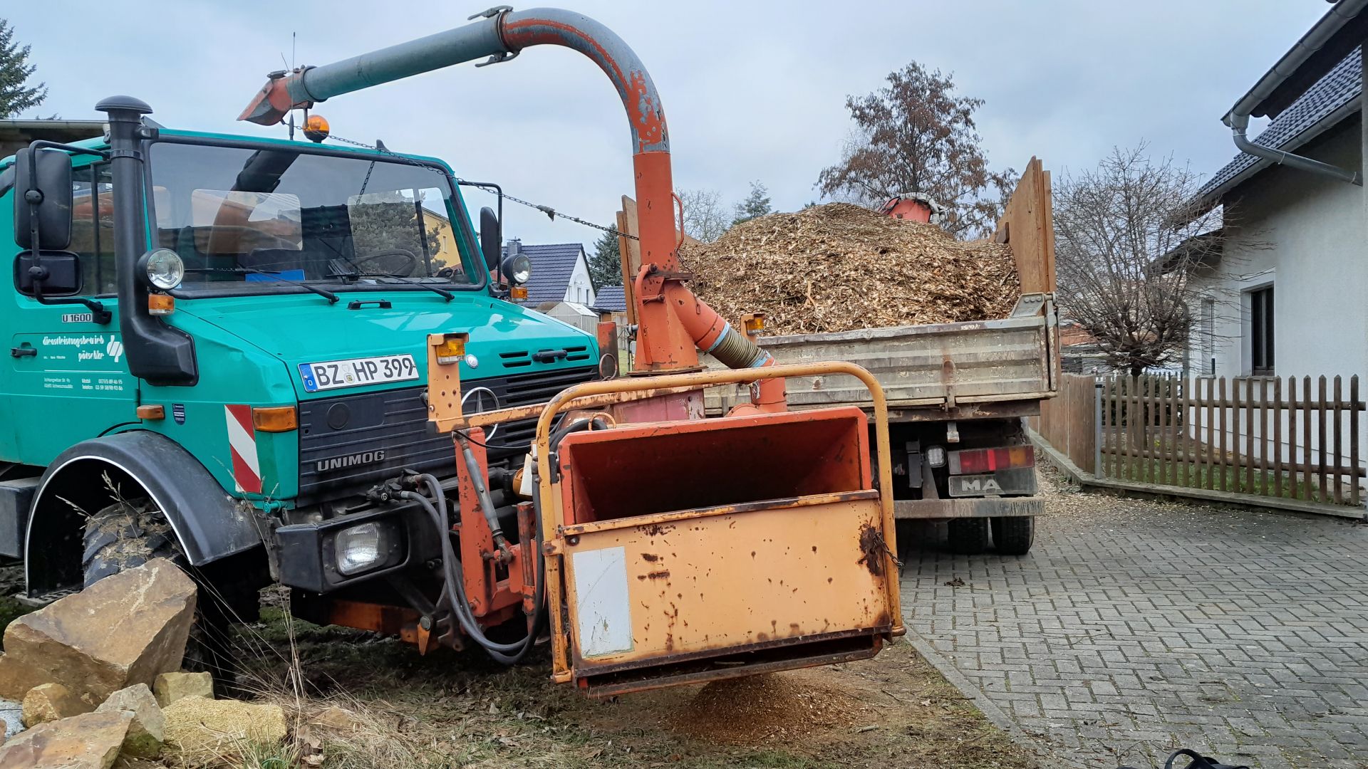 Ein grüner Unimog mit Holzhäcksler verarbeitet Baumverschnitt zu Hackschnitzeln, die in einen Anhänger geblasen werden.