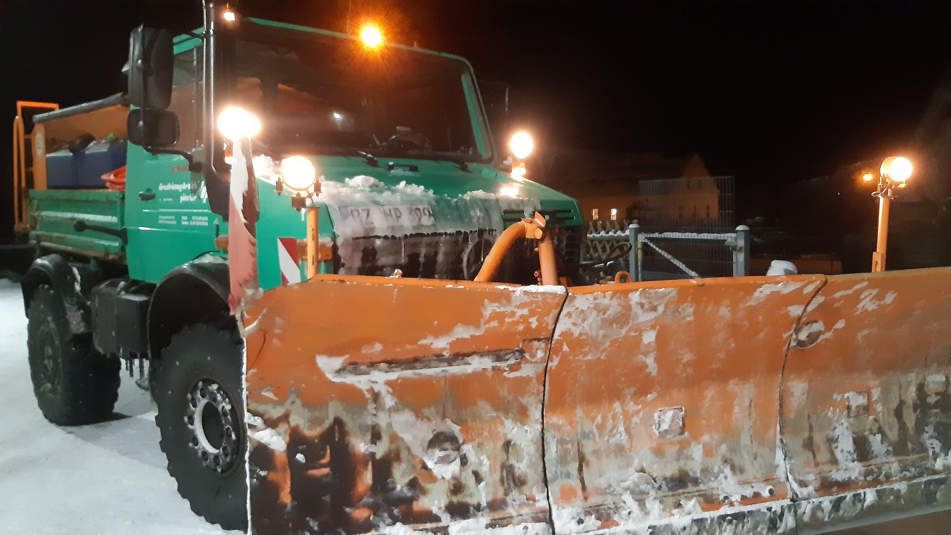 Ein grüner Unimog mit montiertem, orangefarbenem Schneepflug im Einsatz bei Nacht im Winterdienst.