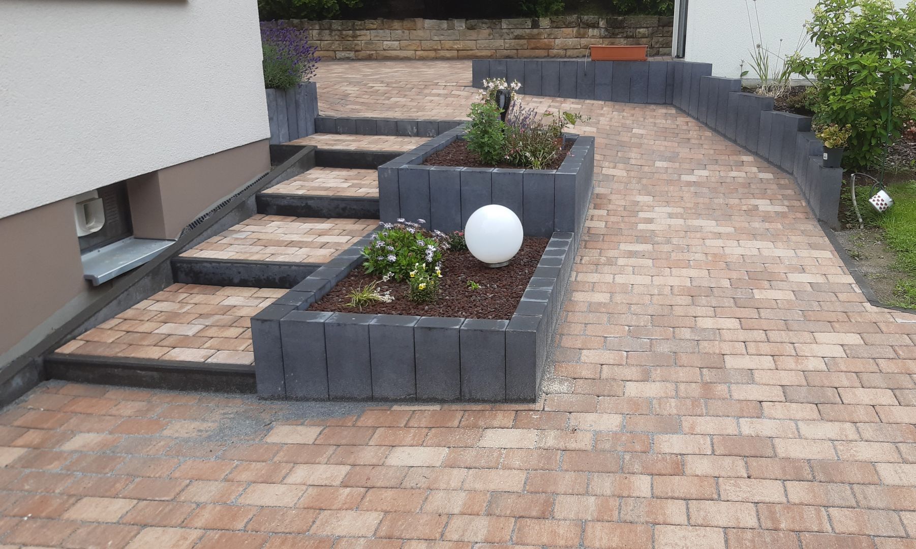 Gestaltete Außenanlage mit rotbraunem Pflaster, Treppe, grauen Hochbeeten mit Pflanzen und einer weißen Gartenkugel.