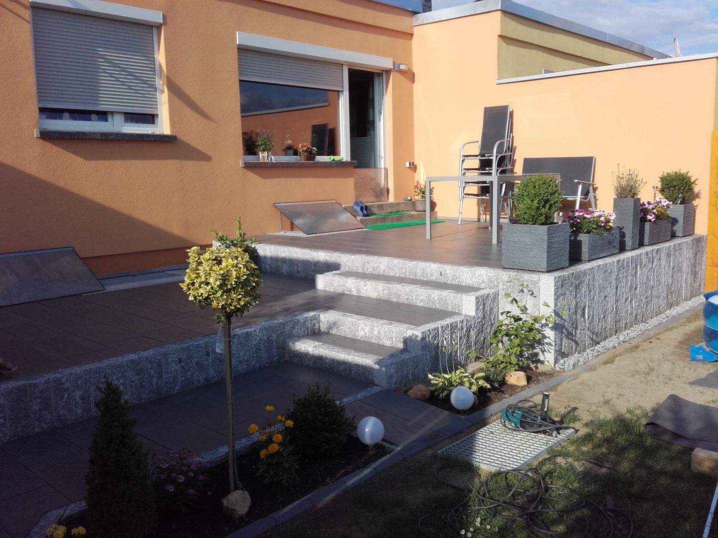 Mehrstufige Terrasse mit dunklem Pflaster und hellgrauen Natursteinmauern, Sitzgruppe und Bepflanzung.