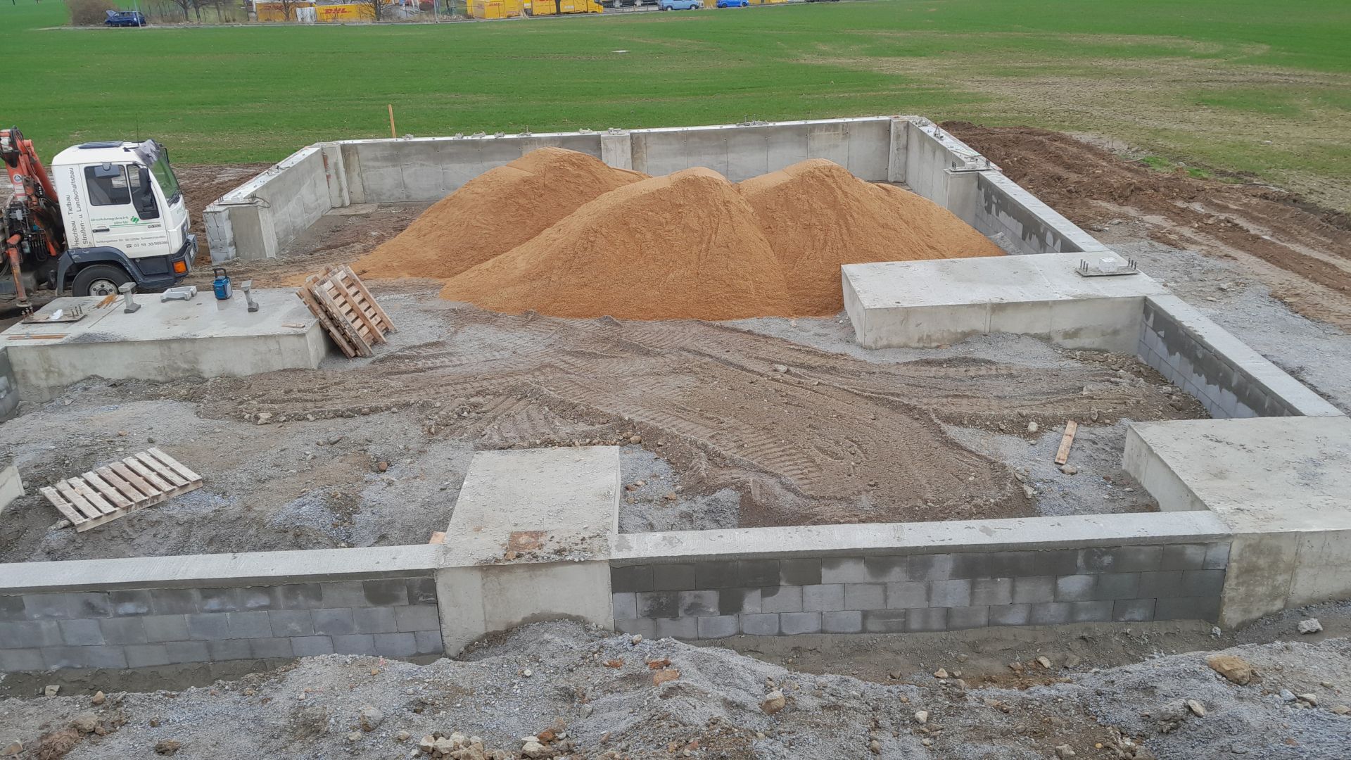 Fundament und Kellerrohbau eines Hauses, mit Betonmauern und Sandhaufen im Inneren der Baugrube.