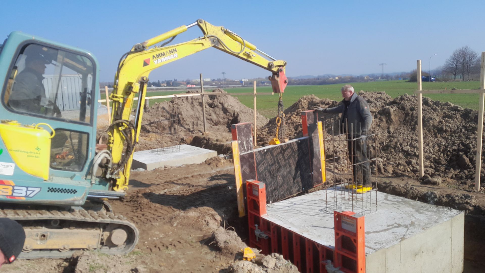 Ein gelber Minibagger hebt ein Schalungselement für den Kanalbau, ein Arbeiter steht in der Baugrube.